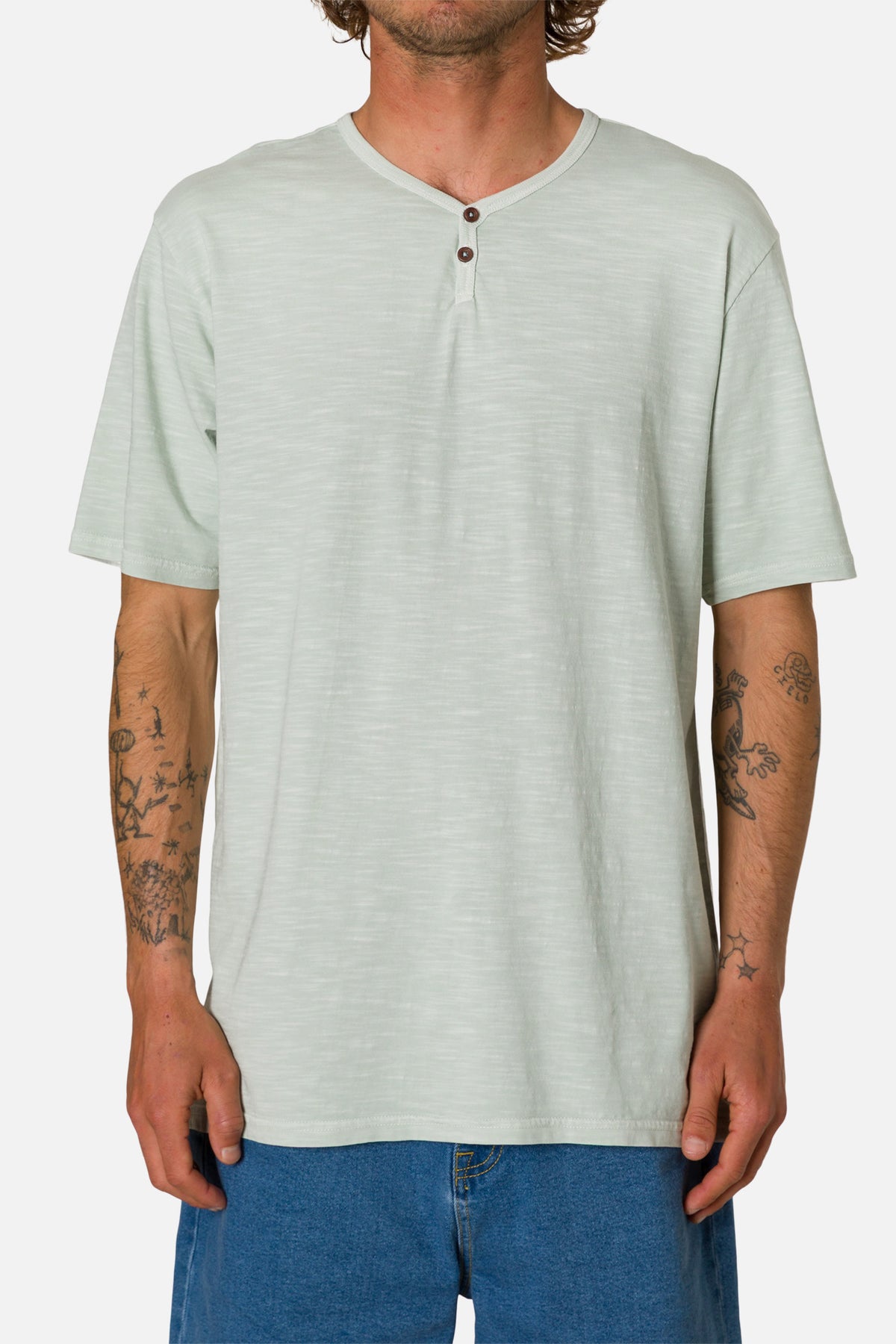 Man wearing a light green henley shirt and blue jeans on a white background