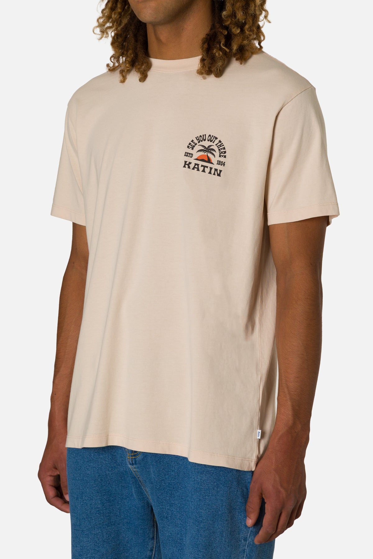 Person wearing a beige t-shirt with a logo and text, standing against a white background