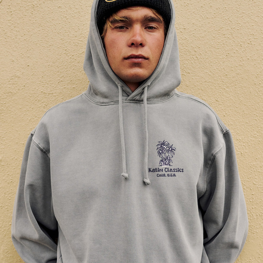 Man wearing a gray Katin Classics palm-tree hoodie and black beanie against a beige wall.