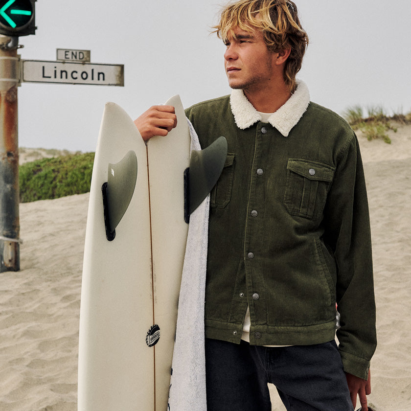 Man wearing an olive corduroy jacket with a white fleece collar while holding a surfboard at the beach.