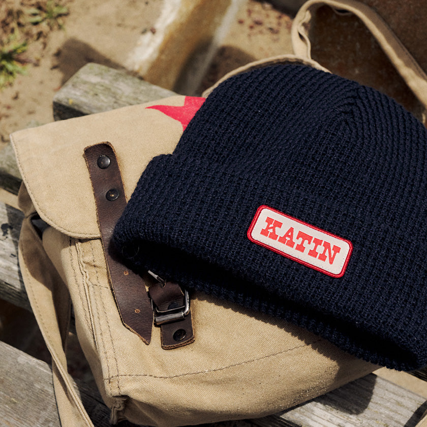Navy ribbed beanie with a red-and-cream Katin patch resting on a tan canvas bag.