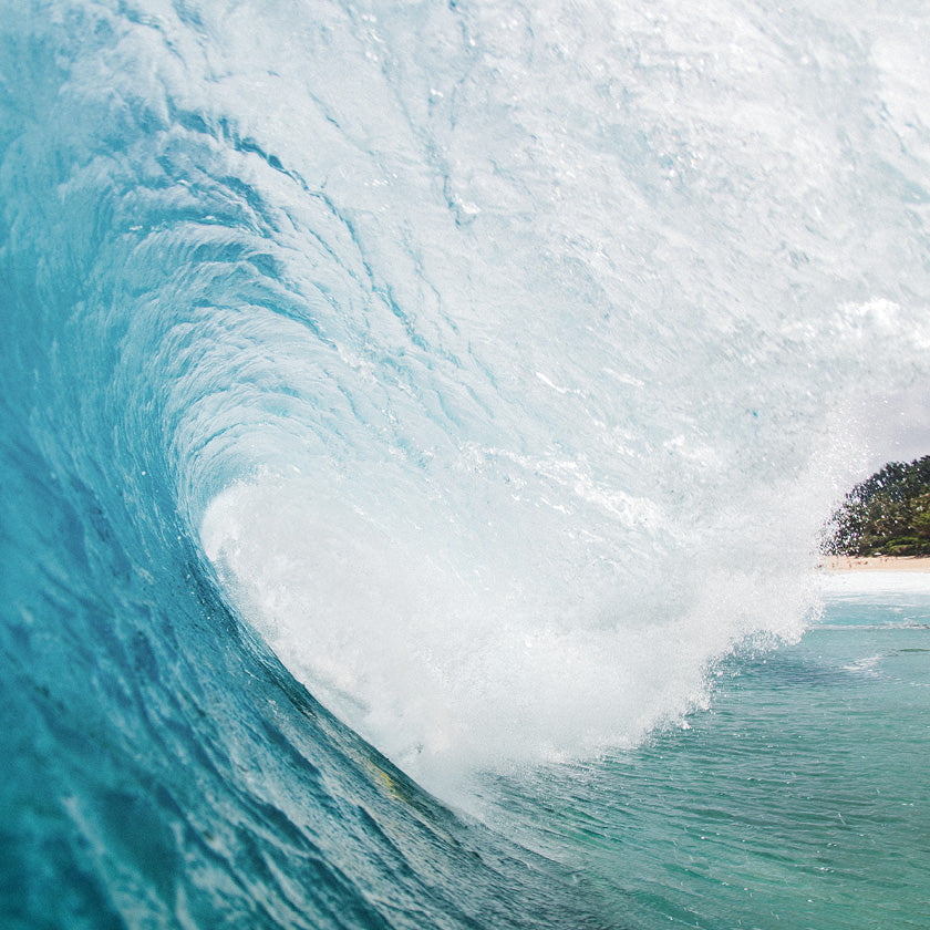 Close-up inside a curling turquoise wave with white spray and a glimpse of a sandy beach.