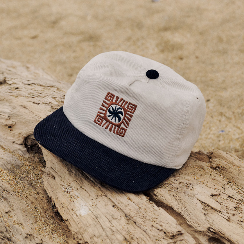 Cream corduroy cap with a navy brim and embroidered palm design resting on driftwood.