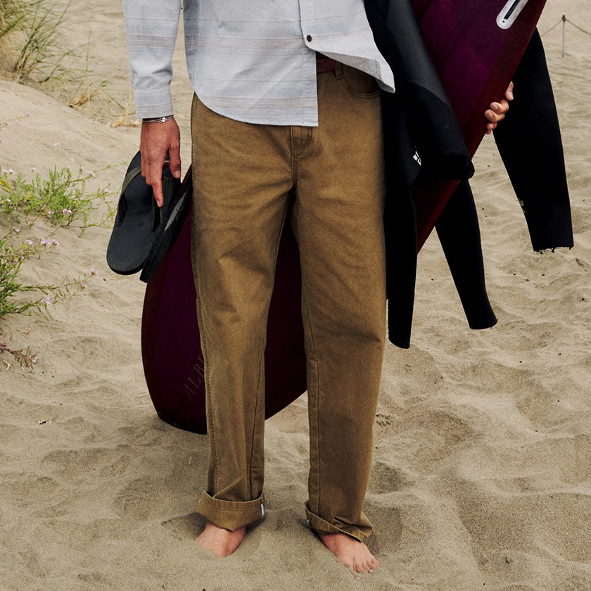 Brown cuffed pants worn barefoot on the sand while carrying a surfboard, wetsuit, and sandals.