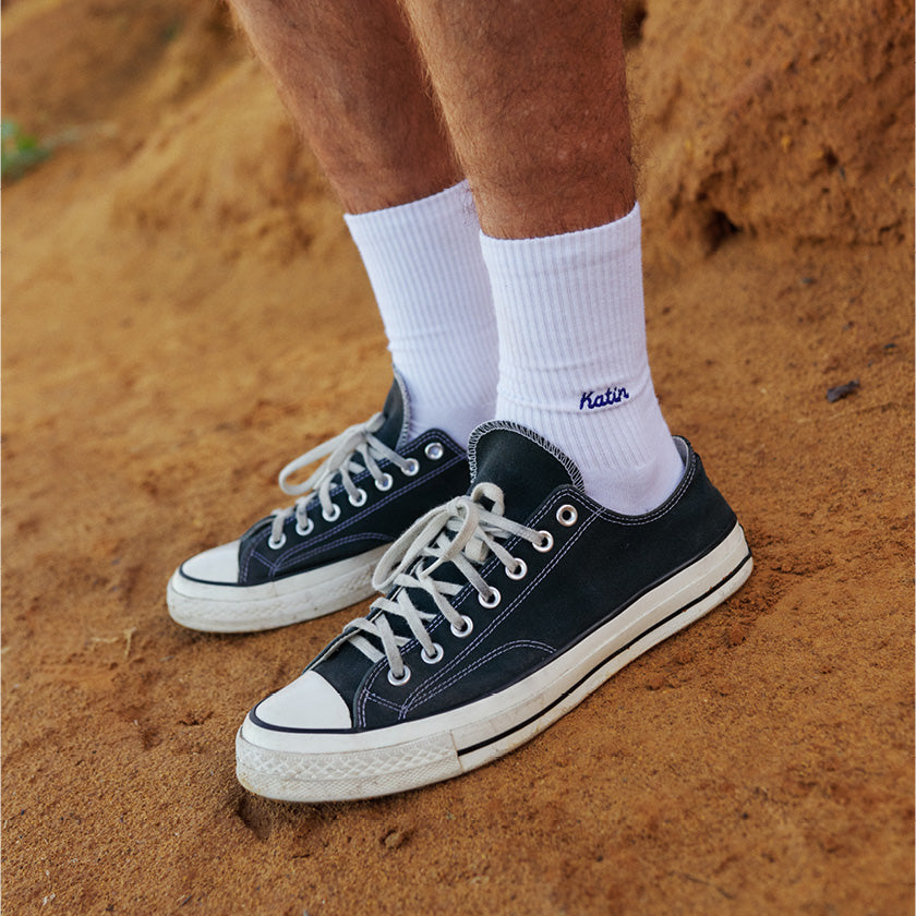 White Katin crew socks worn with black canvas sneakers on a sandy path.