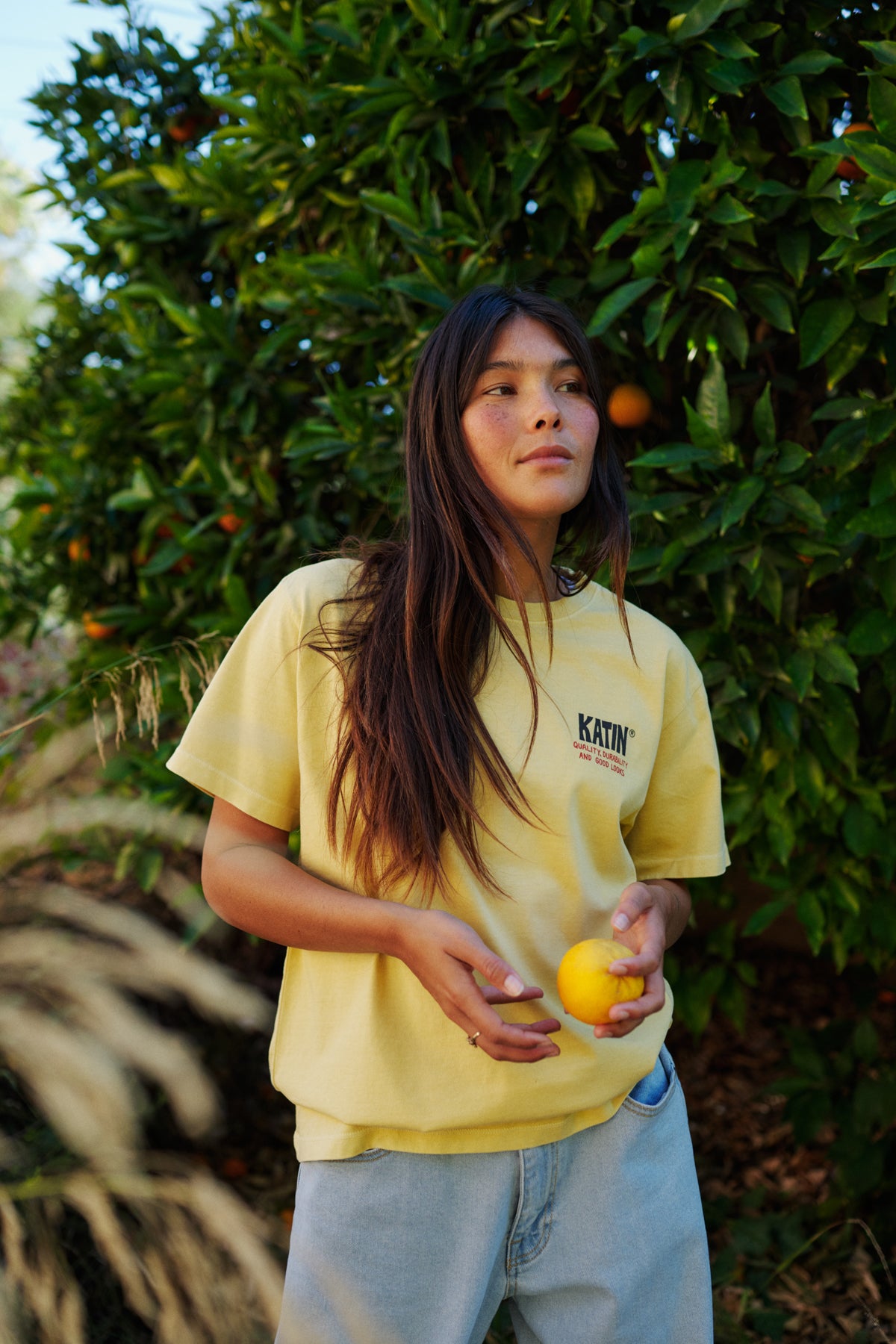 Person holding a orange in front of an orange tree wearing a yellow katin tee