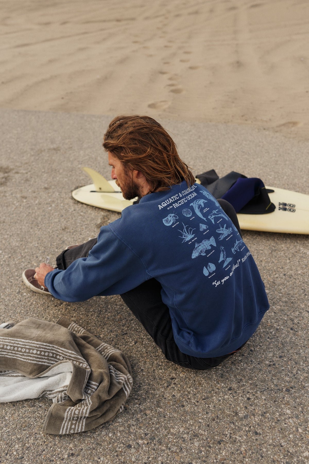 Man sitting on a beach with surfboards, wearing a blue sweatshirt with graphics.