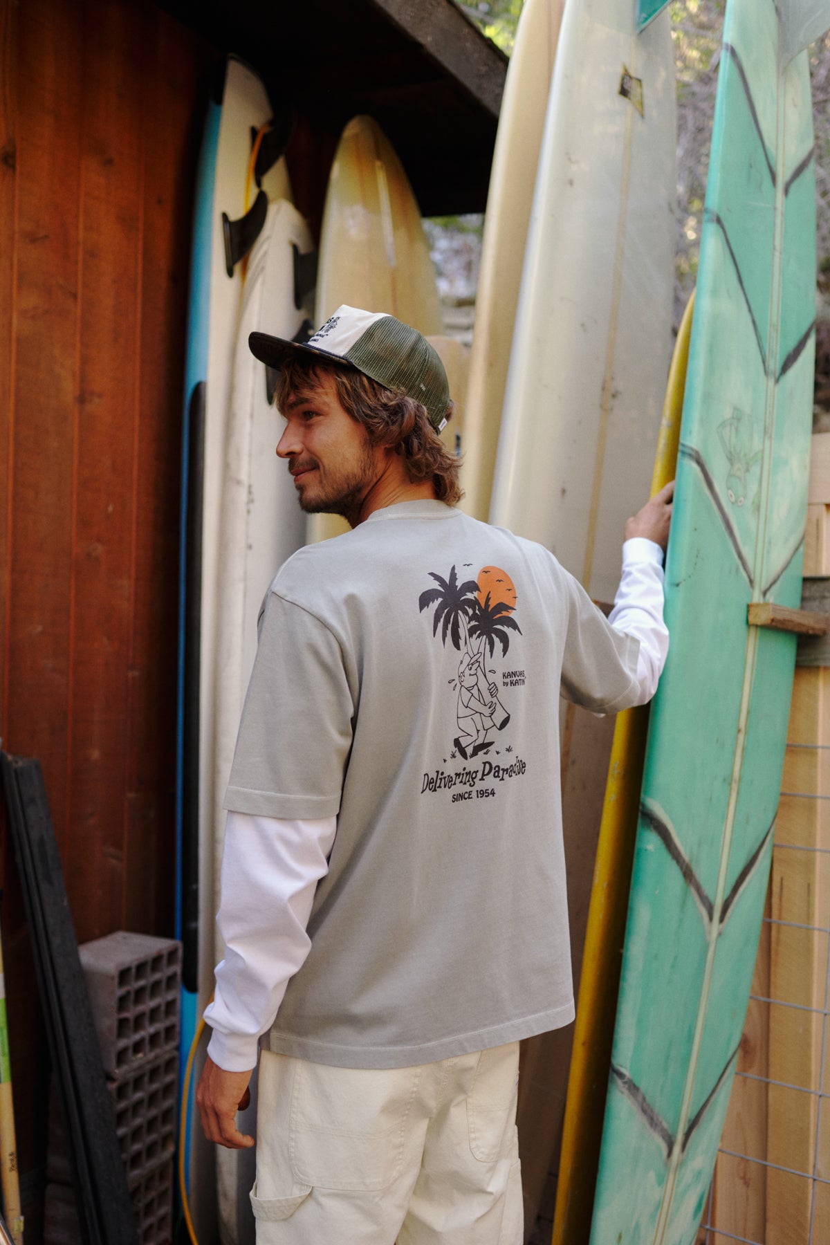 Man holding surfboards with a graphic t-shirt, standing in front of a wooden wall.
