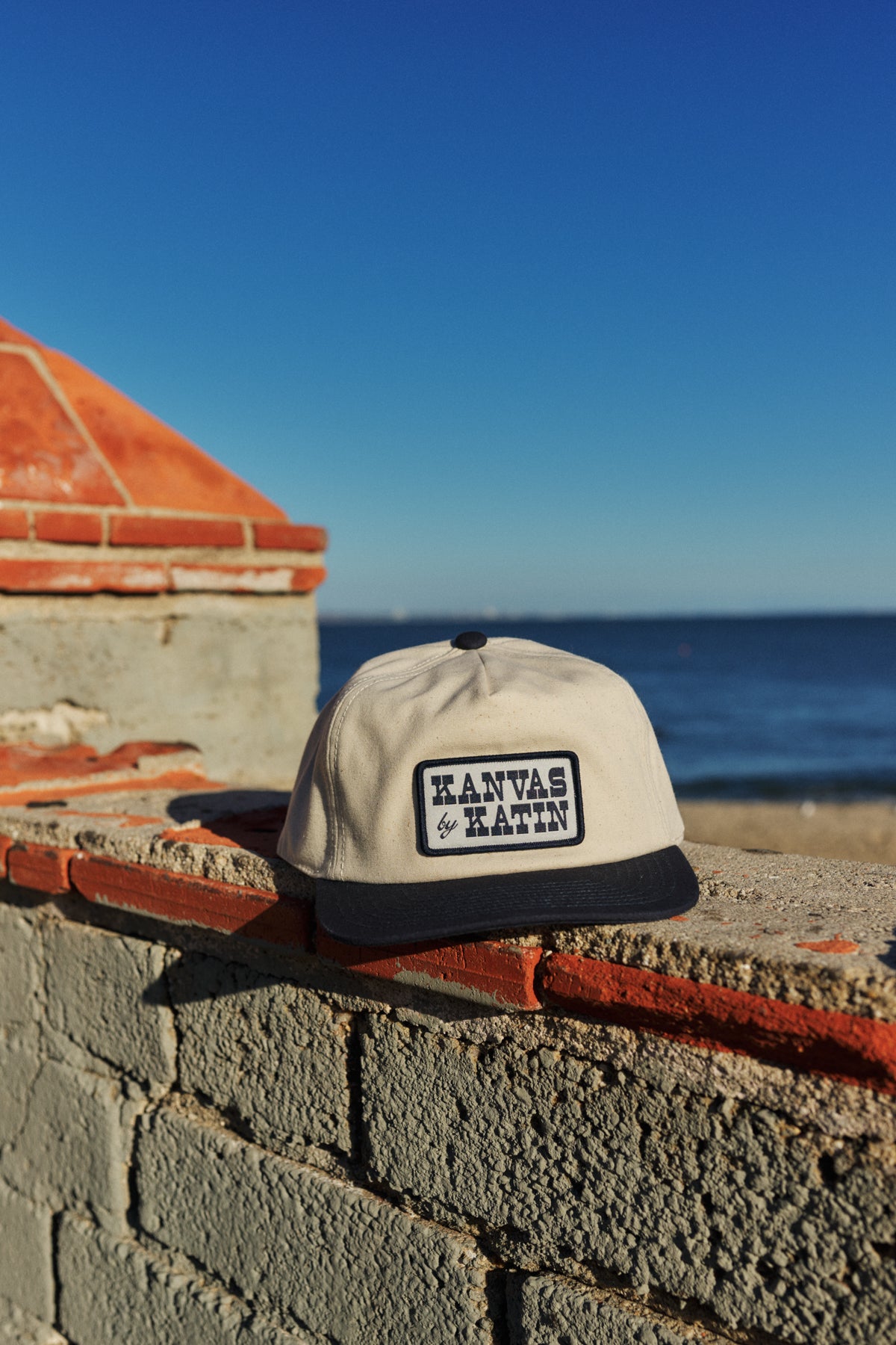 Cap with 'Kanvas & Katin' branding on a brick ledge by the ocean.