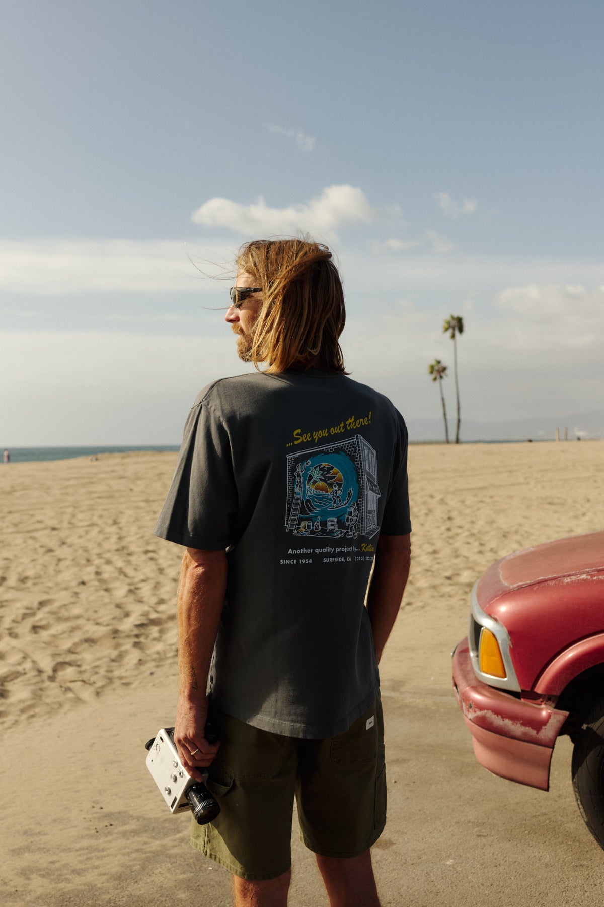 Man standing on a beach with a red truck, wearing sunglasses and a graphic t-shirt.