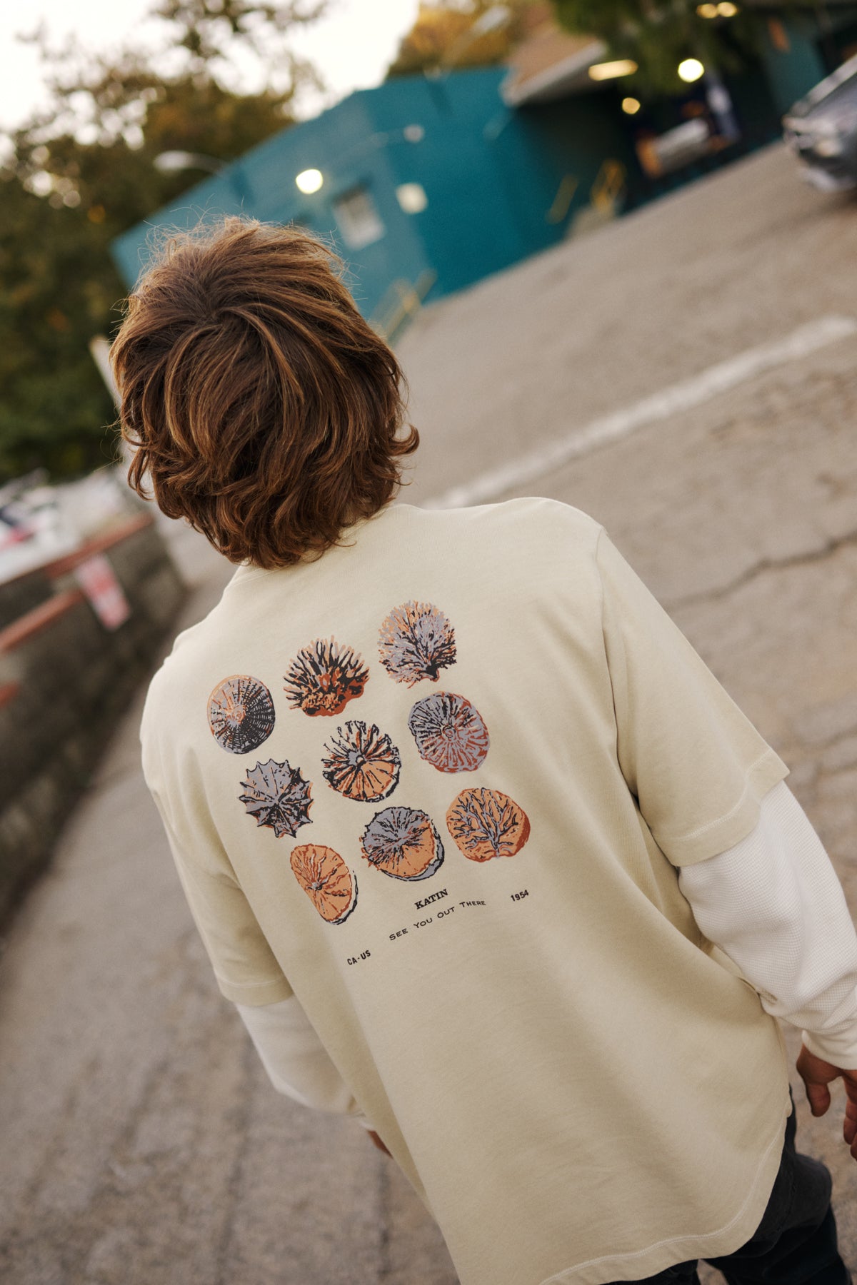 Person wearing a beige shirt with colorful designs on the back, walking on a street.