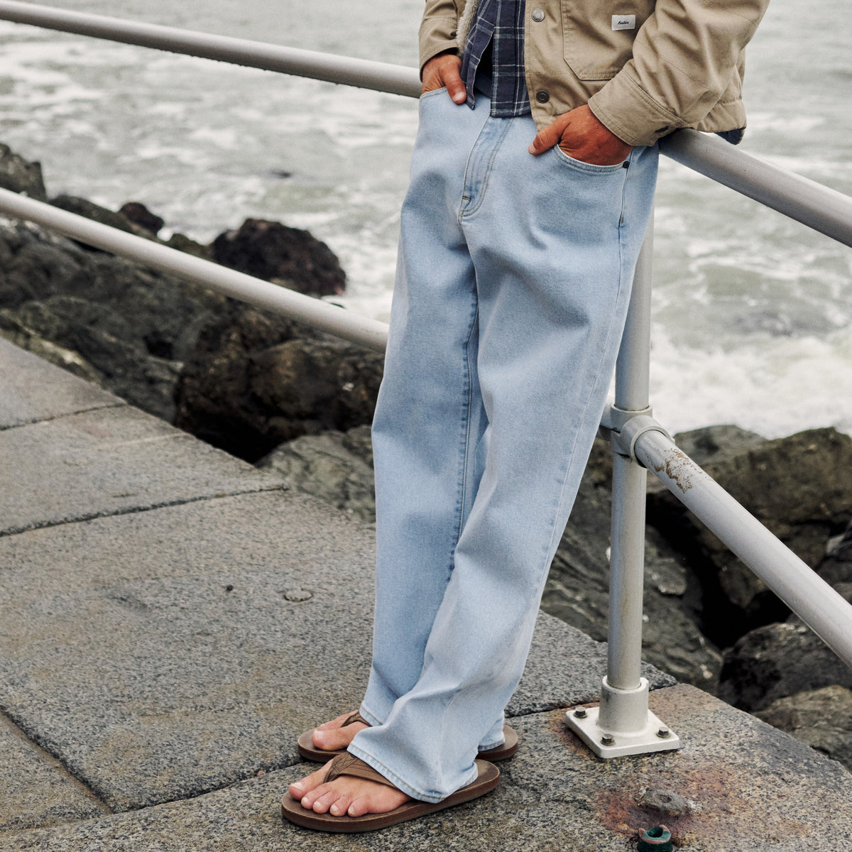 Light-wash jeans worn with a beige jacket and flip-flops beside an oceanfront railing.