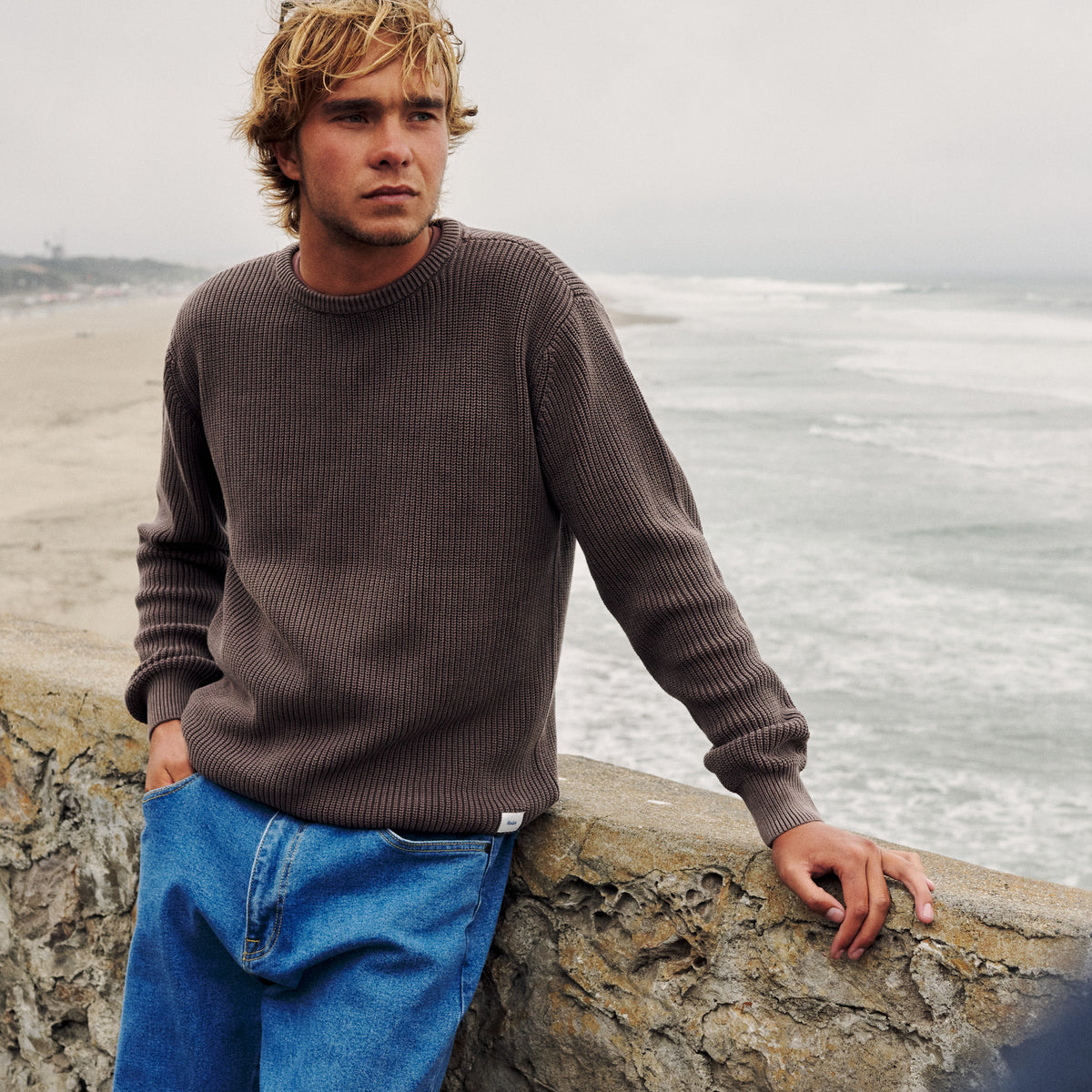 Man wearing a brown ribbed crewneck sweater and blue jeans beside a beachside stone wall.