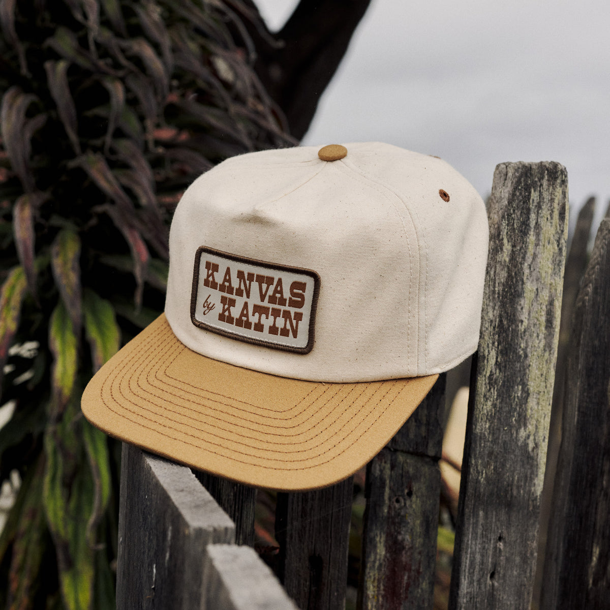 Field Hat in Vintage White/Ermine, a canvas five-panel with a Kanvas by Katin patch, resting on a weathered wooden fence