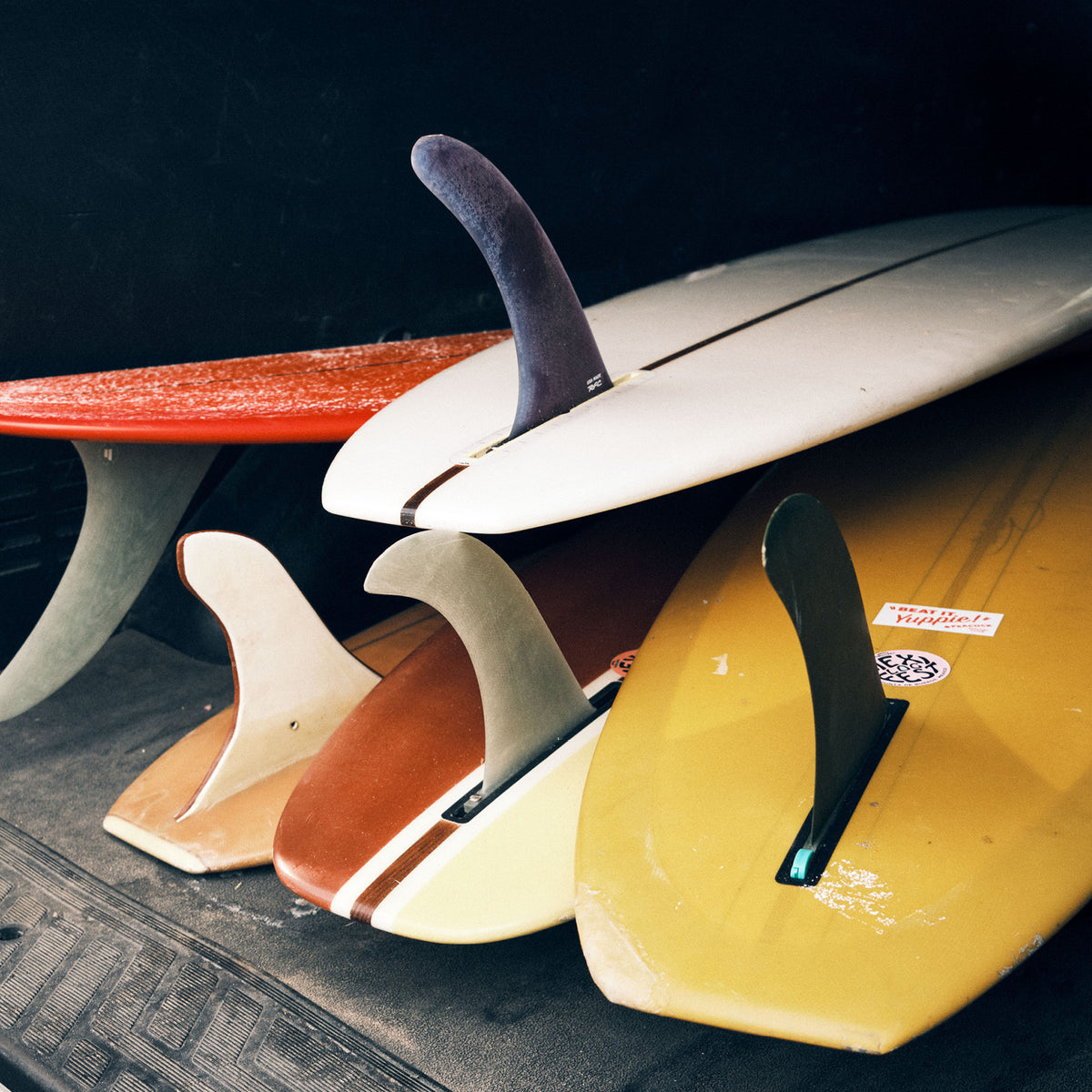 Tails and fins of a row of vintage longboards in red, white, brown and yellow, stacked in the back of a truck