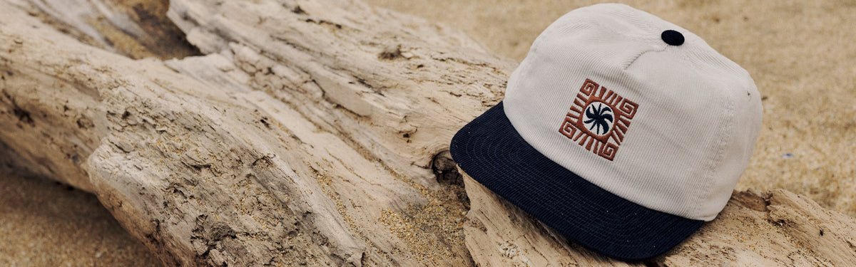 Cream corduroy five-panel hat with a navy brim and an embroidered sun and palm patch, resting on driftwood in the sand