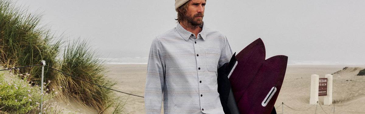Man in the Stevie Shirt in Light Blue, a fine-wale striped corduroy button-up, carrying a surfboard and wetsuit at Ocean Beach