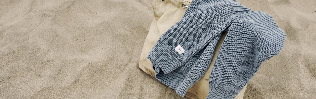 Swell Sweater in Tempest Blue, a ribbed cotton knit, draped over a canvas bag in the sand