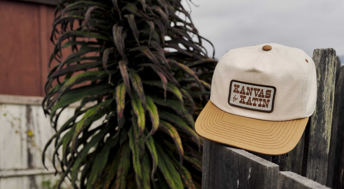 Field Hat in Vintage White/Ermine, a canvas five-panel with a tan brim and Kanvas by Katin patch, resting on a weathered fence beside a coastal plant