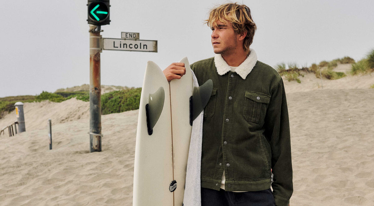 Man in an olive jacket with a white fleece collar holding a surfboard beside a beach traffic light.