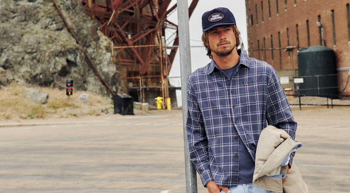 Man leaning against a sign post wearing a navy plaid overshirt and a 'Katin Customs' trucker hat, holding a tan corduroy jacket, with a rocky hillside and industrial ironwork structure behind him.