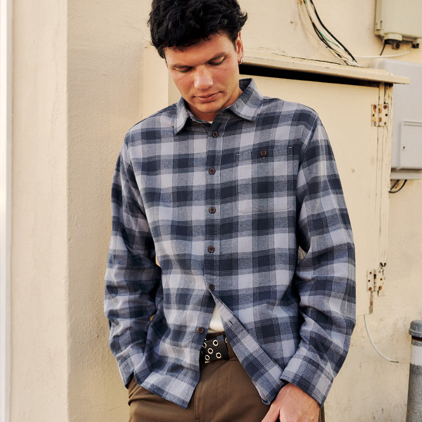 Man wearing a gray-and-navy plaid button-up shirt with brown pants beside a cream wall.