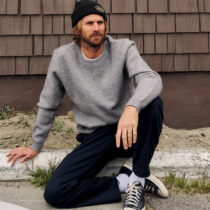Man in a gray crewneck sweater, black beanie, and dark pants sitting beside wood siding.