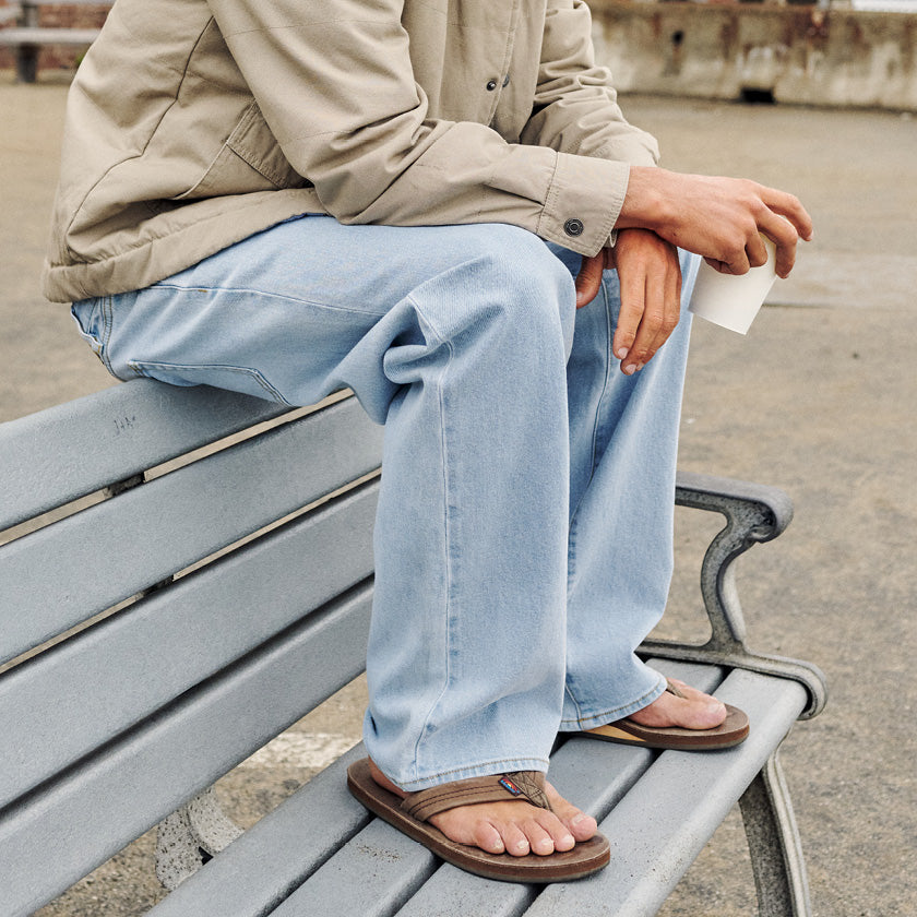 Light-wash jeans and flip-flops worn by a man sitting on a bench in a beige jacket.