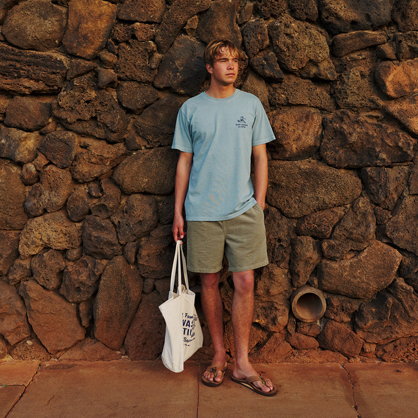 Man in olive shorts and a light blue graphic T-shirt holding a tote bag beside a stone wall.