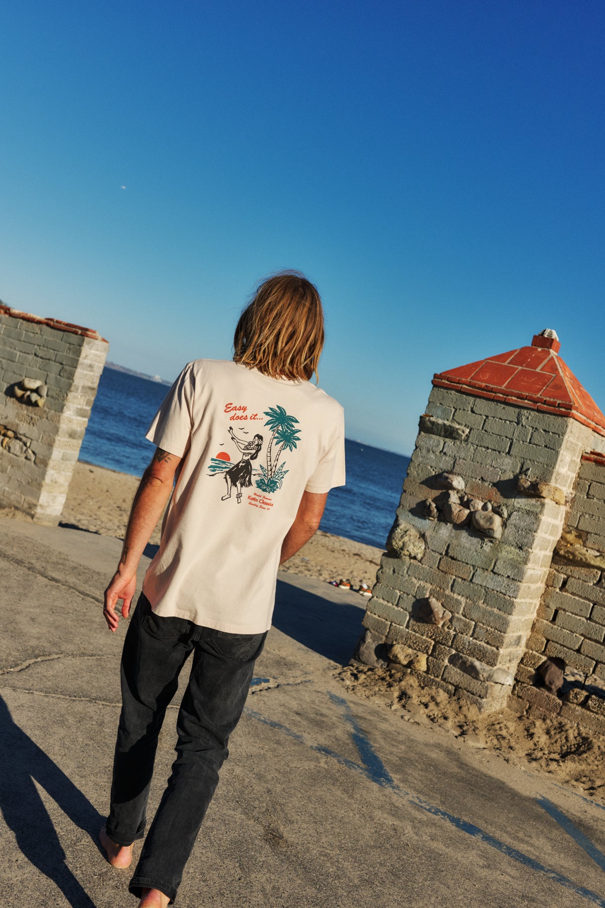 Person walking on a path by a body of water with stone structures and clear blue sky.