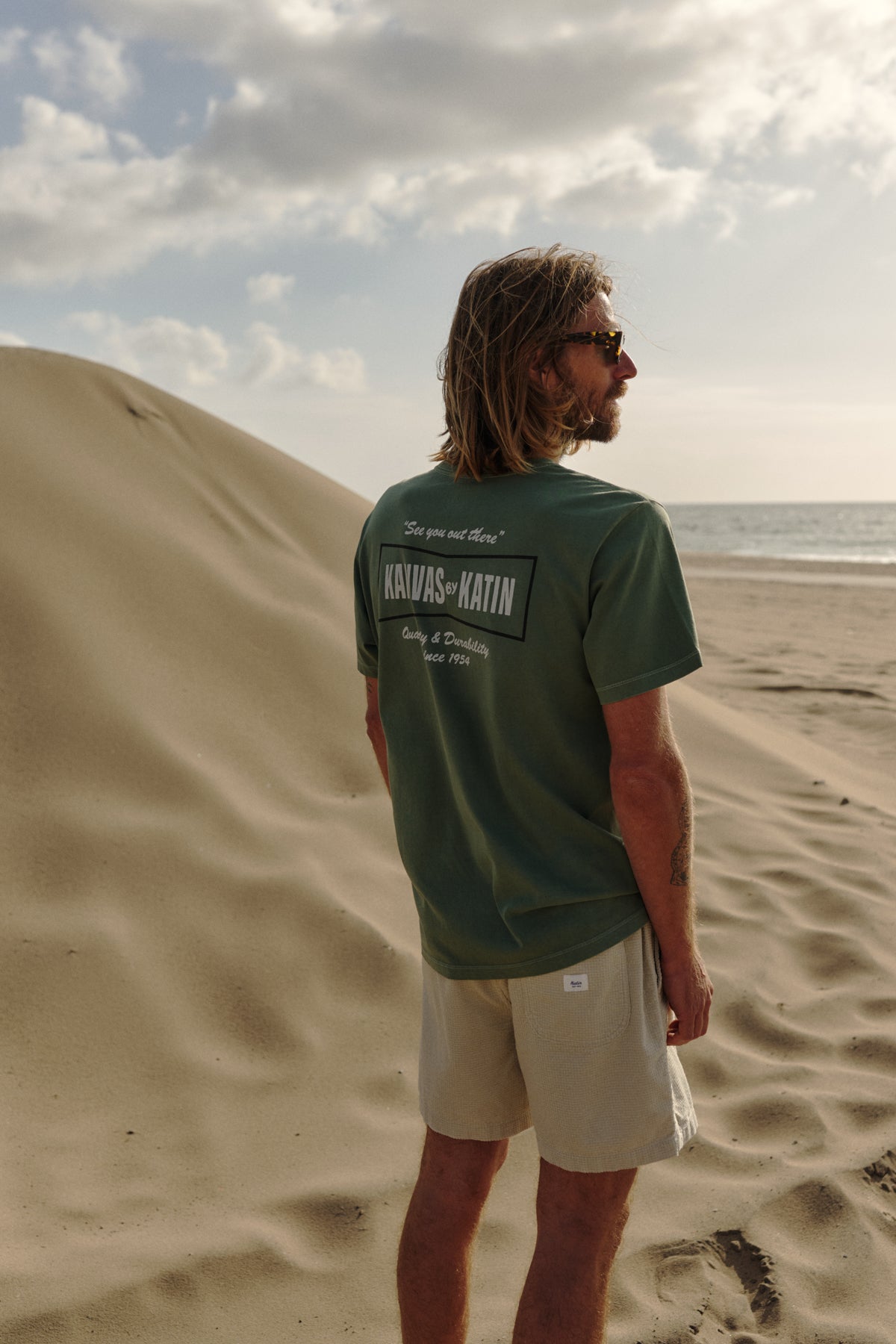 Man standing on a sandy beach wearing a green t-shirt with text on the back.