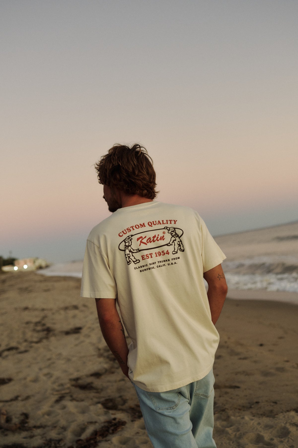 Man standing on a beach wearing a beige t-shirt with text and graphics.