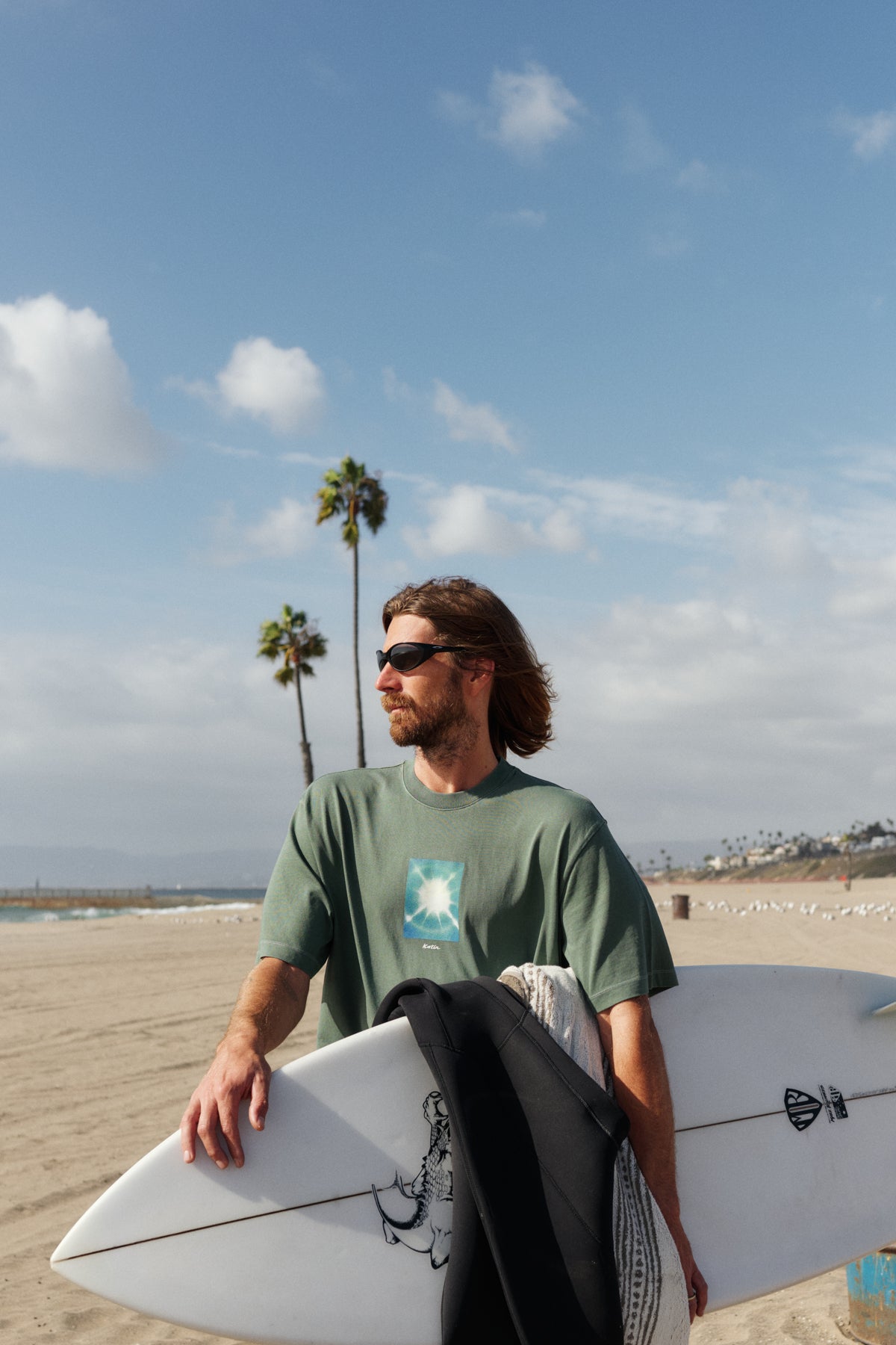Man holding a surfboard on a beach with palm trees and a clear sky.