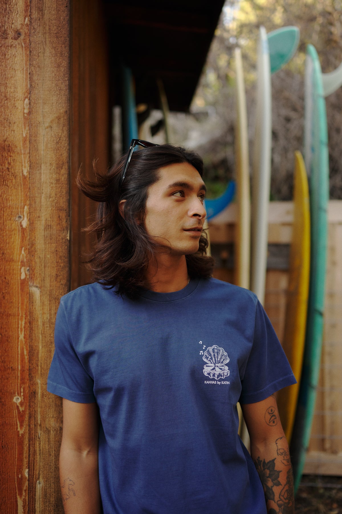 Man wearing a blue t-shirt with a logo, standing in front of surfboards.