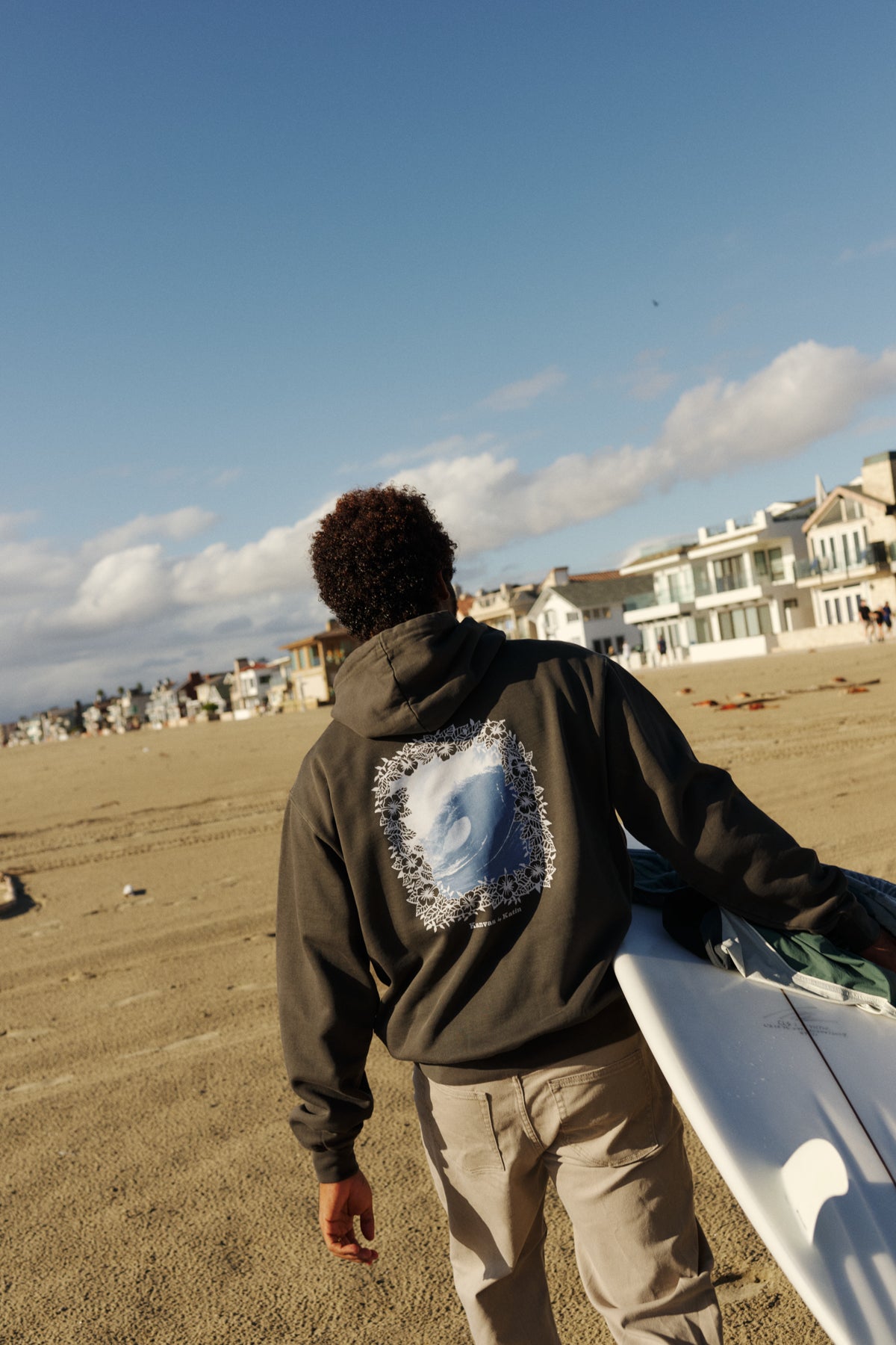 Person walking on a beach with a surfboard, wearing a hoodie with a graphic design.