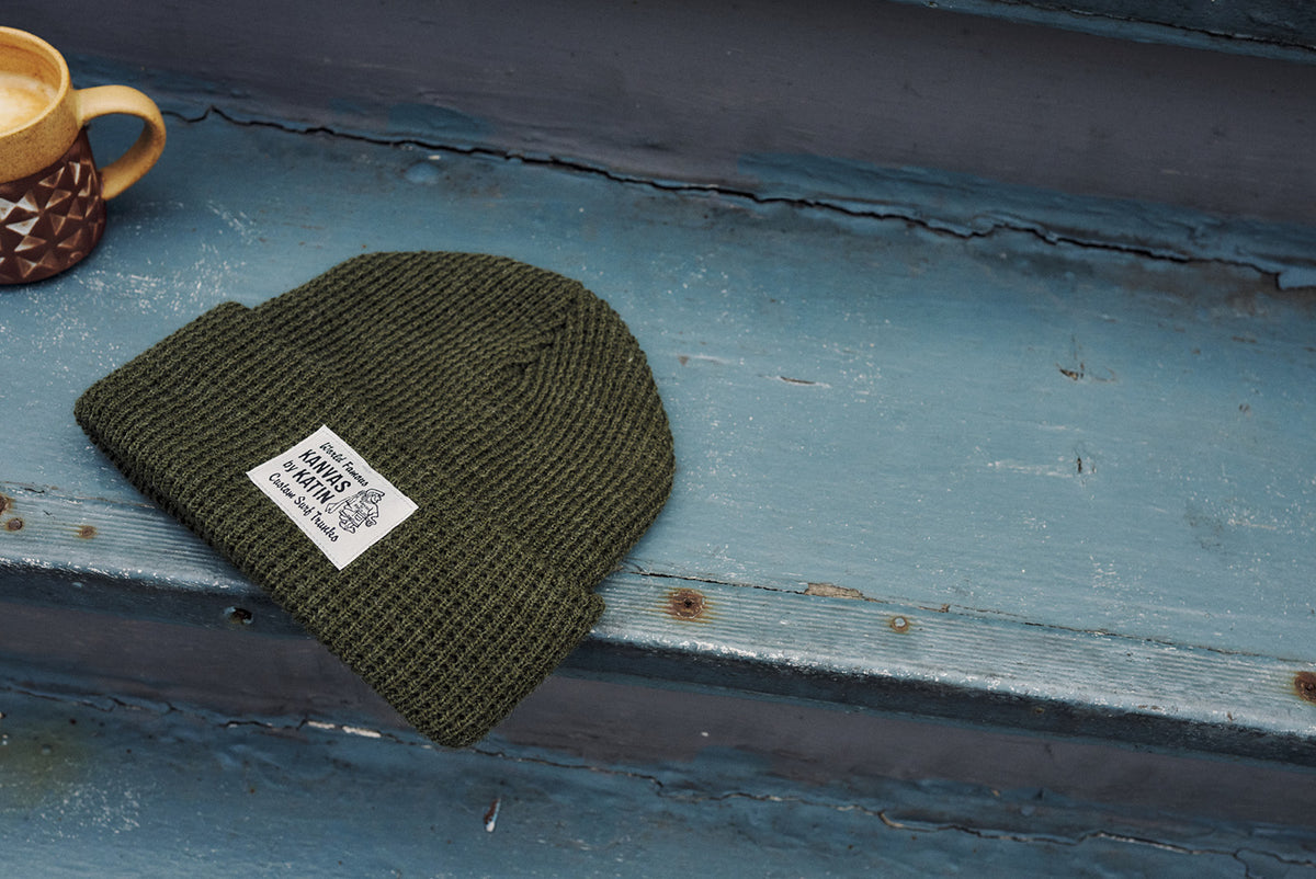 Olive waffle-knit beanie with a “World Famous Kanvas by Katin – Custom Surf Trunks” woven label, on worn blue-painted steps beside a coffee mug