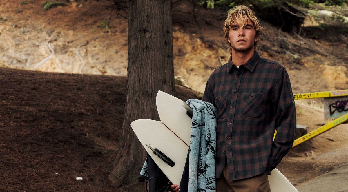 Surfer in a brown and black check Sierra Flannel, carrying a surfboard and palm-print towel past a pine tree and dirt hillside