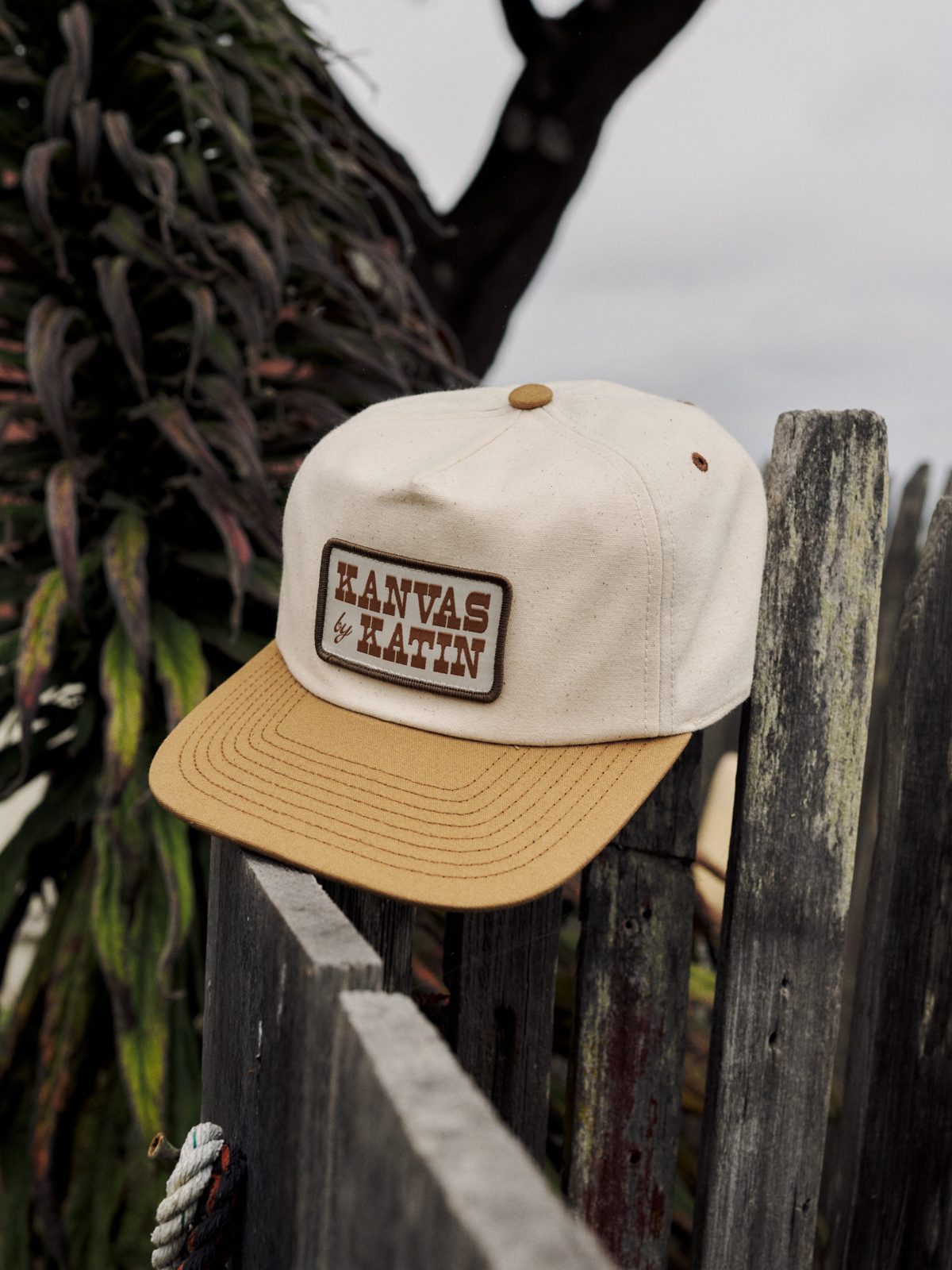 Katin Field Hat in Vintage White/Ermine, cotton canvas five-panel with a Kanvas by Katin patch, on a weathered fence, lifestyle #color_vintage white/ermine