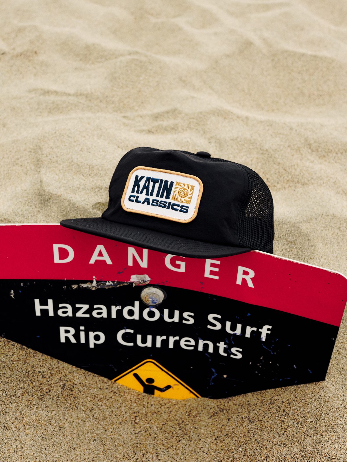 Katin Helios Trucker Hat in Black, recycled nylon with mesh back and Katin Classics patch, on a beach sign in the sand, lifestyle #color_black