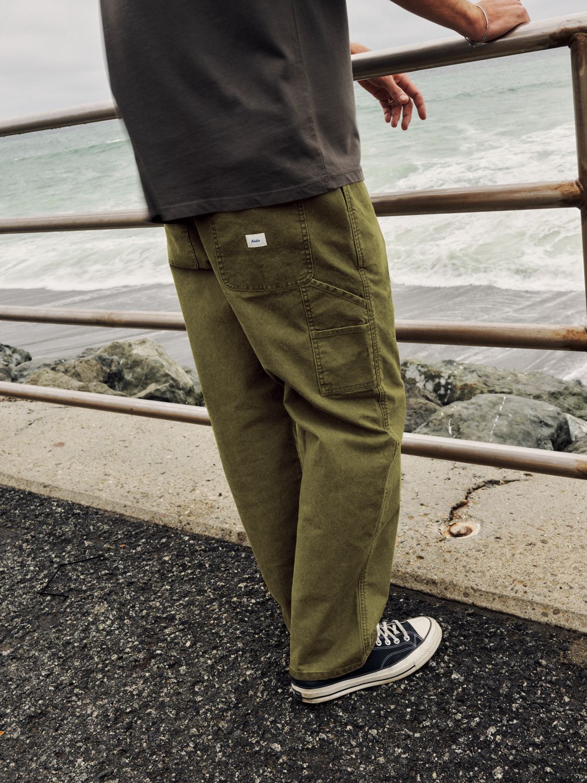 Katin Stanley Pant in Olive, relaxed-fit cotton canvas carpenter pant, on model by the ocean, back view, lifestyle #color_olive